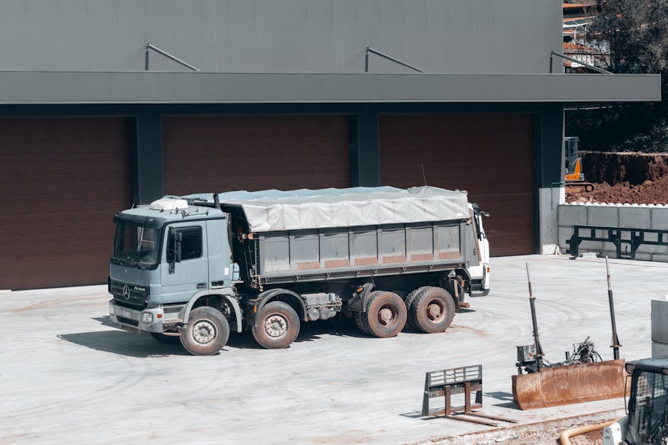 A large light grey Mercedes-Benz truck with a covered cargo area parked on a paved surface in front of a modern building with wooden paneling and grey exterior walls. The truck is positioned near a do