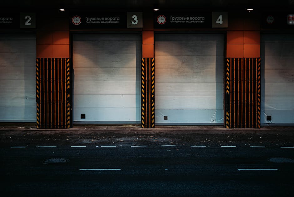 A large light grey Mercedes-Benz truck with a covered cargo area parked on a paved surface in front of a modern building with wooden paneling and grey exterior walls. The truck is positioned near a do