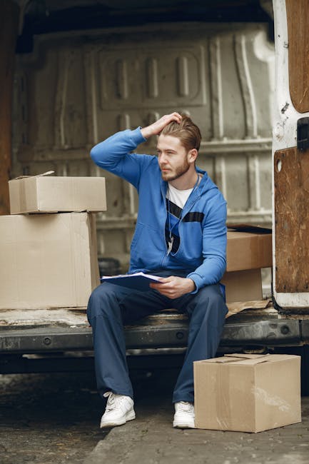 A man wearing a white t-shirt, blue jeans, and a black cap is seated at the rear of an open black van, which is loaded with numerous medium-sized and smaller cardboard boxes, some stacked on top of ea