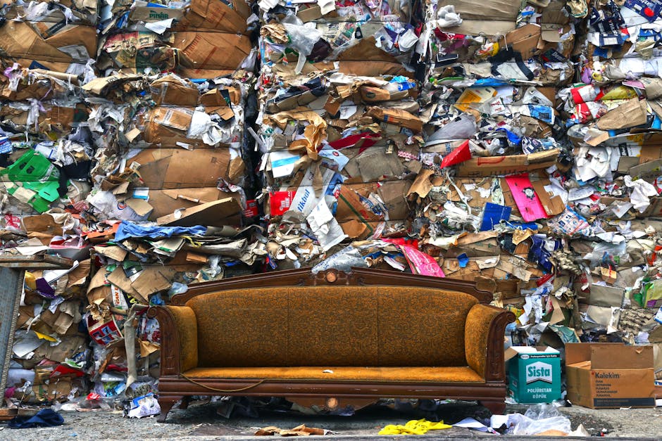 A vintage brown upholstered sofa with carved wooden armrests and backrest is positioned against a wall of piled cardboard boxes and crumpled paper, many sealed with tape, and assorted packaging materials in a chaotic heap. The sofa appears to be outdoors or in an area prepared for loading, situated near a doorway or pavement, indicating it may be in the process of being moved or disposed of. Nearby, a small white box labeled 'Sista' and a brown cardboard box can be seen on the ground, with visible lighting suggesting daytime. This scene exemplifies packing and loading activities involved in home relocation or furniture transport, with Man with Van Northwood likely handling such bulky waste removal or moving logistics.