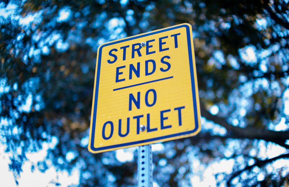 A close-up photograph of a rectangular yellow street sign with bold blue text reading 'STREET ENDS NO OUTLET', mounted on a metal pole with numerous holes for adjustable height. The sign is positioned outdoors, with a background of blurred dark green foliage and branches of trees under natural daylight. The sign is clean and clearly visible, indicating the end of a street without an outlet, which is relevant for relocation or home moving logistics in Northwood. The image captures the sign's reflective surface and the environmental setting typical for vehicular and pedestrian navigation, supporting the context of street closures in house removals or transportation planning as handled by Man with Van Northwood.
