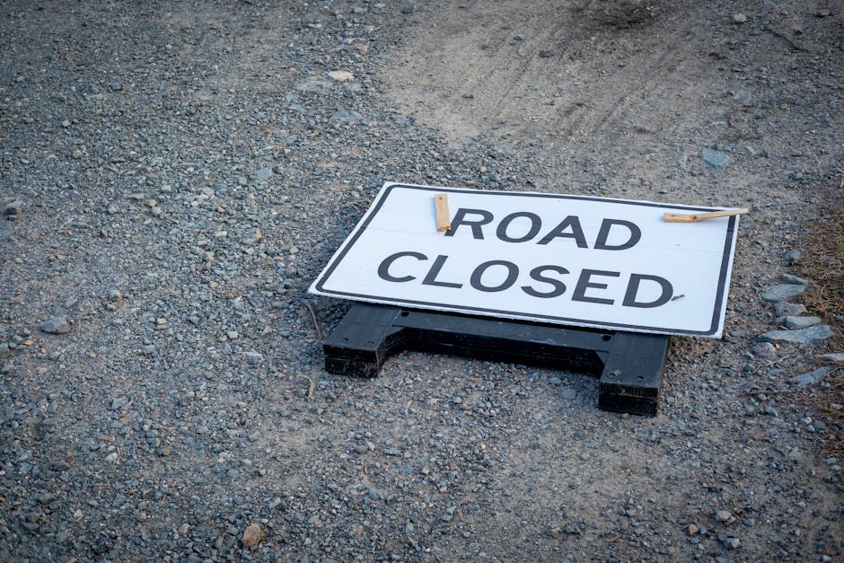 A black and white 'Road Closed' sign with bold black lettering is positioned on the gravel ground outside a property, supported by two wooden clothespins clipped onto a horizontal string. The sign is placed near the entrance, indicating that the road access is temporarily unavailable, likely due to ongoing home relocation or moving activities. The area surrounding the sign consists of loose gravel and small stones, suggesting an outdoor setting, possibly a driveway or street nearby. In the background, the lower part of a building’s wall or pavement is faintly visible, with no other objects or vehicles in the immediate vicinity. The setup is indicative of a professional removals service, such as Man with Van Northwood, managing a residential move that may require road closure permits or temporary access restrictions during furniture transport and packing processes. The sign’s positioning highlights the importance of coordinating logistics for efficient home relocation within Northwood.