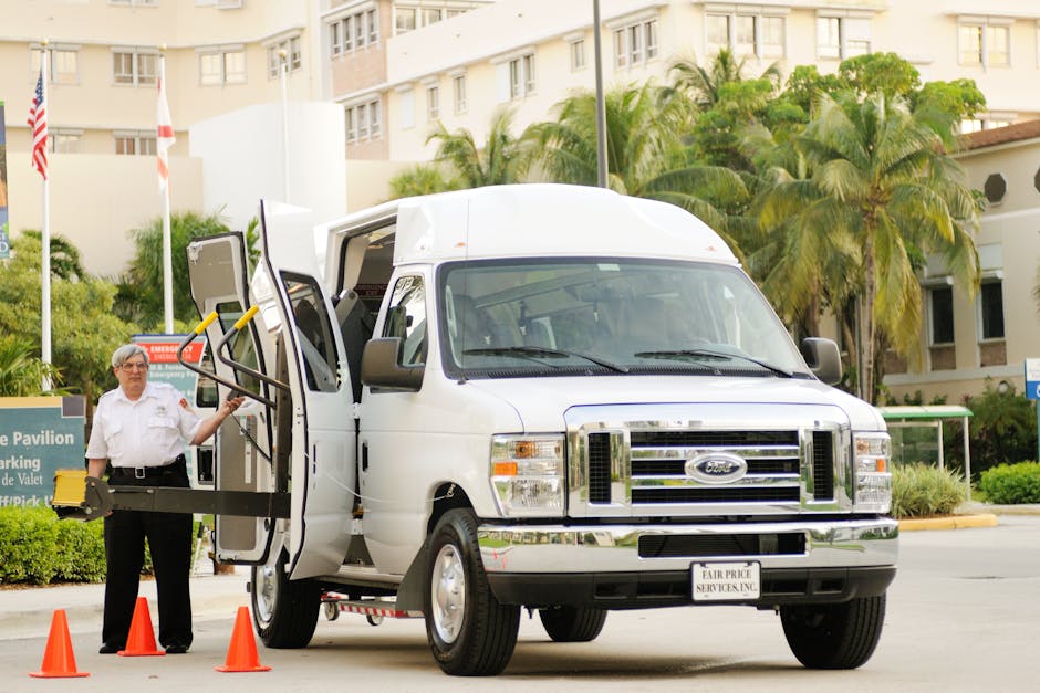 A man with grey hair and glasses wearing a white shirt and black trousers stands next to a white Ford transit van, which is parked in a residential area with tall palm trees and apartment buildings in the background. The van doors are open, revealing the interior used for home relocation or furniture transport. The man is holding onto a set of yellow-handled safety straps or handrails attached to the side of the van, which are secured on a black platform or loading ramp. Orange traffic cones are placed around the loading area to ensure safety during the loading process. Inside the open van, parts of packing materials such as cardboard boxes, plastic wrap, and possibly wrapped furniture are visible, indicating a move-in or move-out operation. The environment is well-lit, with clear weather, illustrating a typical house removal or moving service scenario undertaken by Man with Van Northwood, emphasizing their process of packing, loading, and transport of household items through narrow roads or urban settings.