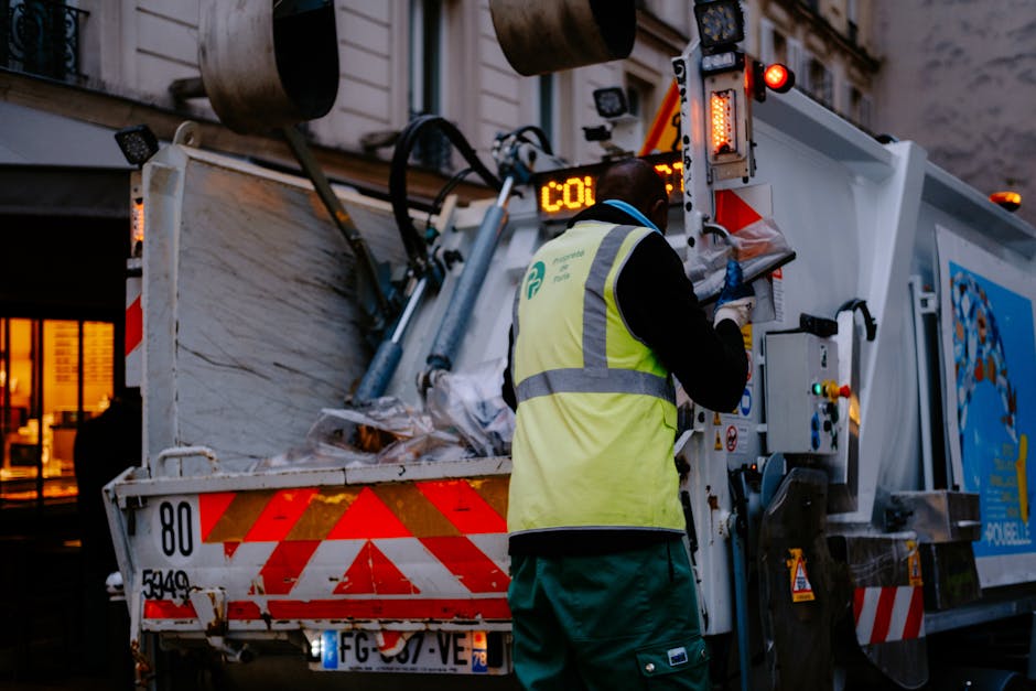 A worker from Man with Van Northwood, wearing a high-visibility yellow vest with reflective strips and dark clothing, is operating a waste collection vehicle during dusk or early evening. The worker is standing beside the rear of the bin lorry, which is partially loaded with various waste materials, including cardboard boxes and plastic packaging, some of which are visibly crumpled or flattened. The vehicle features red and white chevron reflective markings on its rear, as well as red warning lights illuminated at the top. The scene takes place on a street with illuminated buildings in the background, indicating urban surroundings. The worker appears to be checking or recording details on a control panel situated on the side of the waste vehicle, which is part of a home removal or clearance process. This image highlights the logistics involved in waste disposal during house removals within Northwood, aligning with the services provided by Man with Van Northwood, especially in compliance with local waste rules for house relocations.