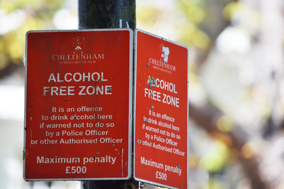 A close-up photograph of two red Cheltenham Borough Council 'Alcohol Free Zone' street signs mounted on a black metal pole outdoors, with a blurred background of trees and foliage. The signs feature white text warning that drinking alcohol in this area is an offence, with a maximum penalty of £500, and note that the offence can be enforced by police or other authorised officers. The signs show some minor wear and peeling paint, indicating exposure to the elements. The outdoor environment suggests a residential or public area, commonly associated with community safety signage. While the image is not directly related to house removals, it illustrates typical signage that might be encountered during home relocation or moving logistics, where awareness of local regulations is important. Man with Van Northwood occasionally references such signage in the context of moving and local area regulations.