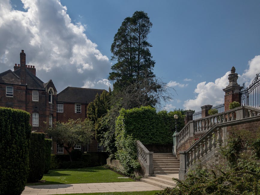 A residential property with red-brick walls, multiple windows, and a pitched roof, situated behind a garden area with neatly trimmed hedges and trees. A wide stone staircase with a decorative balustrade leads up from the paved pathway in the garden to an ornate brick and stone entrance terrace, which is bordered by a cast-iron gate. The garden features a tall tree with sparse foliage, a smaller bushy tree, and well-maintained grassy patches. The sky above is partly cloudy with patches of blue, indicating a bright day. This scene reflects the exterior environment of a house suitable for a professional home relocation or furniture transport service, such as those provided by Man with Van Northwood, with accessible outdoor stairs and a spacious pathway for moving logistics.
