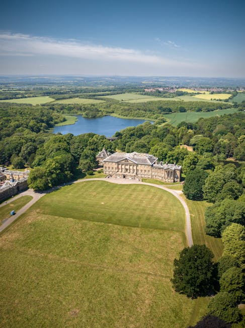 Aerial view of a large historic mansion with classical architectural features, surrounded by extensive green lawns and tall mature trees, situated within a spacious estate. Behind the mansion, there is a small lake bordered by dense woodland, with open fields and farmland extending into the distance under a partly cloudy sky. The scene showcases a serene, rural landscape typical of the Moor Park Estate area, capturing the natural surroundings that may require careful planning during home relocations. The image emphasizes the property's setting, which is relevant to house removals and moving services like those offered by Man with Van Northwood, highlighting the importance of accurate access and logistical considerations for furniture transport and packing during moving processes.