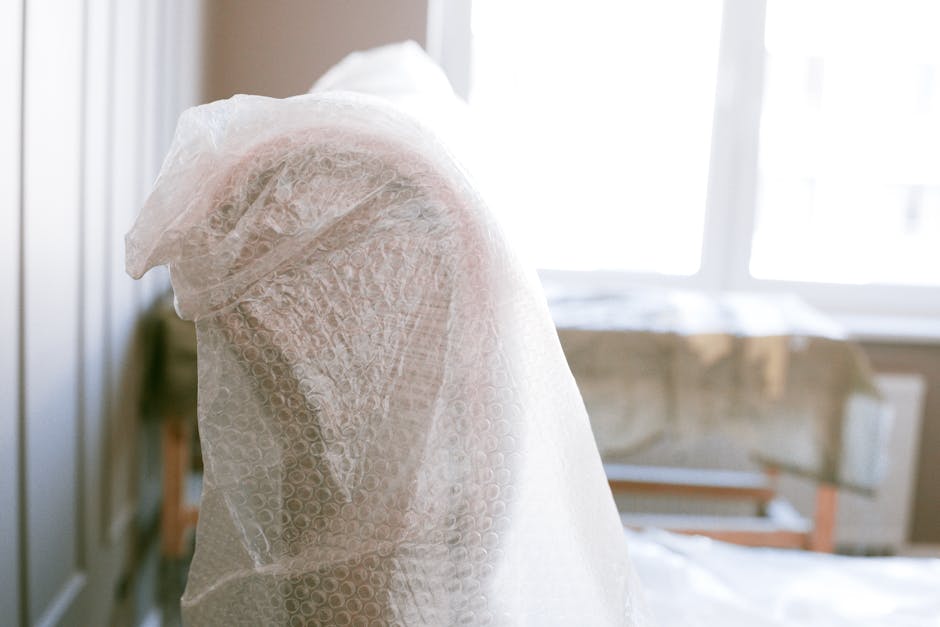 A wrapped piece of furniture, likely a chair or small table, is covered with bubble wrap for protection during home relocation. The item is positioned inside a room near a window, with natural light illuminating the packaging materials. The bubble wrap securely envelops the object, indicating careful packing and moving practices by Man with Van Northwood, specialists in removals and furniture transport. In the background, a partially visible beige couch or another piece of furniture rests on a protective covering, possibly on a moving trolley or clean surface, ready for transportation. The room appears to be in the process of being prepared for a professional move, with items packed for safety and stability during loading, reflecting standard packing and moving procedures associated with house removals. The environment is neat and organized, with a focus on safeguarding belongings for an efficient loading process, supported by the company’s comprehensive services.