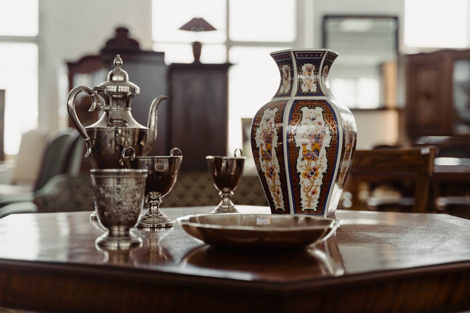A polished wooden dining table inside a well-lit room displays a collection of decorative and functional items, including a tall, ornate ceramic vase with a detailed pattern in shades of blue, orange, and cream, positioned centrally on the table. To the left of the vase, there is a silver teapot with a curved spout and a lid, along with two matching silver cups, reflecting light from the windows. In front of the vase, a shallow metallic bowl is placed. The background shows blurred furniture pieces and large windows that allow natural light to illuminate the space, creating a warm, inviting atmosphere suitable for home relocation and packing logistics. Occasionally, Man with Van Northwood’s removals service may involve handling similar items during the careful packing and furniture transport process, ensuring antique and delicate pieces are secured during house move efforts.