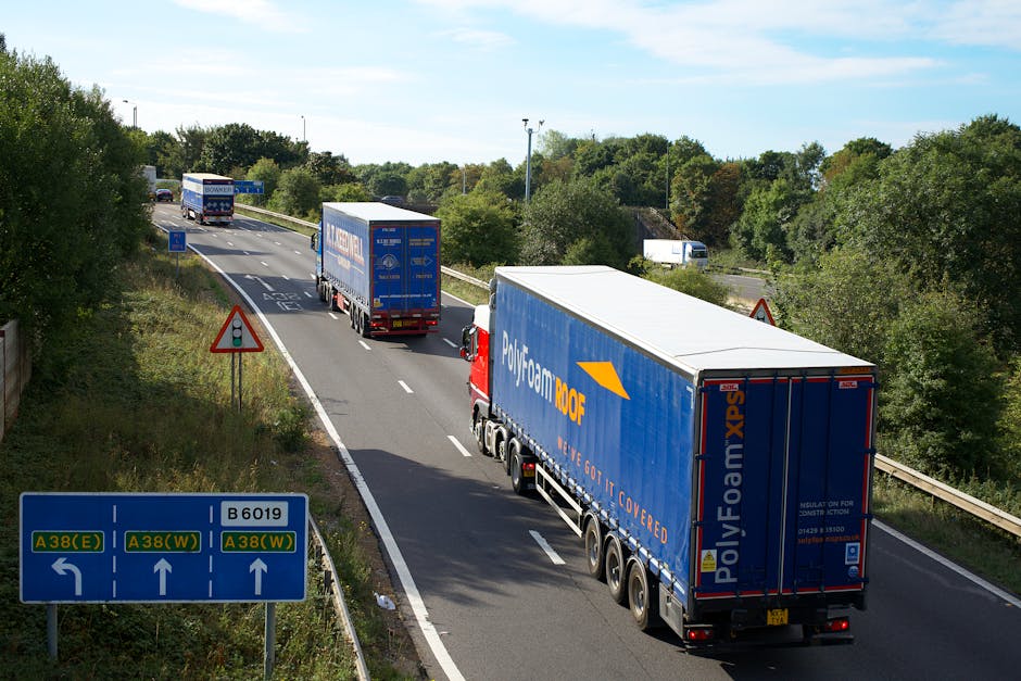 A staged photograph of a highway showing three large trucks transporting furniture and packing materials, possibly during a home relocation or moving process. The trucks are moving along a multi-lane road with a blue directional sign indicating routes A38(E) and A38(W) as well as B6019. The leading truck is a large blue Polyfoam 100F lorry with branding on the side, followed by two more trucks, all in motion. The road is bordered by greenery and trees, with a blue sky overhead. On the left side, there is a warning traffic sign indicating caution for vehicles. The scene captures a typical logistics operation involving furniture transport for house moves, potentially managed by a professional removal service such as Man with Van Northwood, which specialises in house removals and moving services.