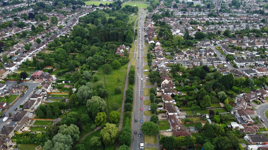 An aerial view of a residential neighbourhood showing a main road running through the centre, lined with parked cars on both sides. To the left of the road, there is a large green park with numerous mature trees and grassy areas, bordered by a narrow footpath. Surrounding the park are rows of detached and semi-detached houses with gardens, fences, and driveways. To the right of the road, there are densely packed houses with small front gardens, some with driveways and parked vehicles. The scene is illuminated by daytime sunlight, highlighting the lush greenery and the various roofing materials of the homes. This image captures a typical suburban setting suitable for house removals, illustrating the environment in which professionals like Man with Van Northwood might facilitate home relocation, furniture transport, and packing and moving services within Northwood and the surrounding areas.