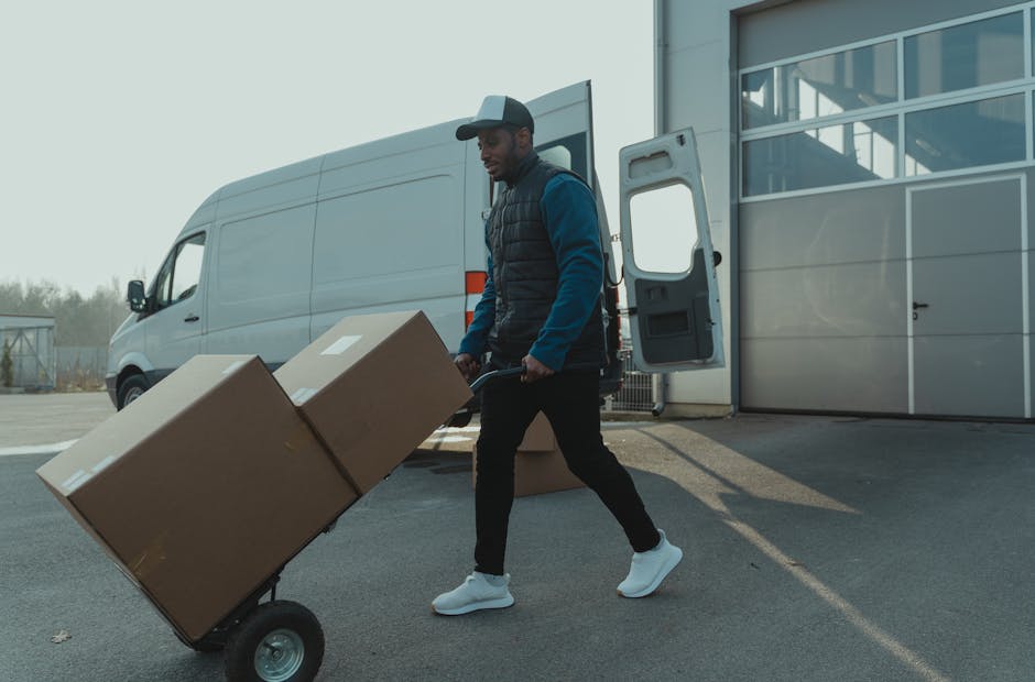 A man wearing a dark baseball cap, black padded vest over a blue long-sleeve shirt, black trousers, and white sneakers is engaged in home relocation activities during daytime. He is walking outside ne